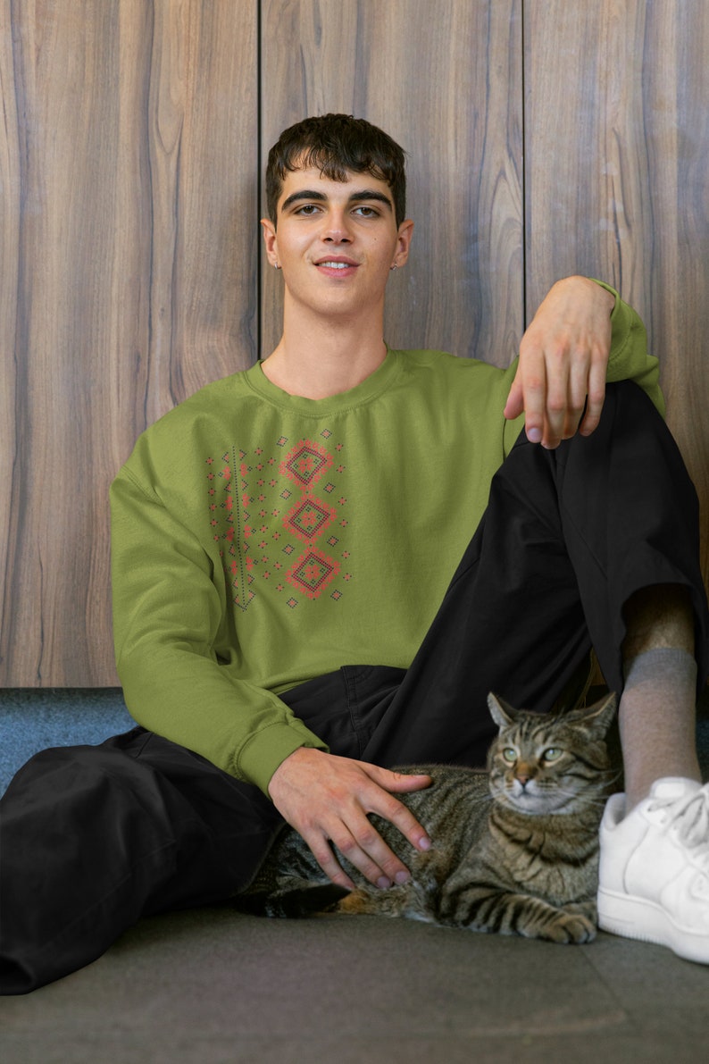 May include: A man wearing a green sweatshirt with a red and black geometric pattern sits on the floor with a tabby cat. He is wearing black pants and white sneakers.