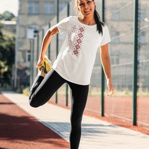 Puede incluir: Camiseta blanca con un dise&ntilde;o geom&eacute;trico en rojo y gris. La camiseta se combina con leggings negros y zapatillas deportivas amarillas. La imagen est&aacute; tomada al aire libre en una pista roja.