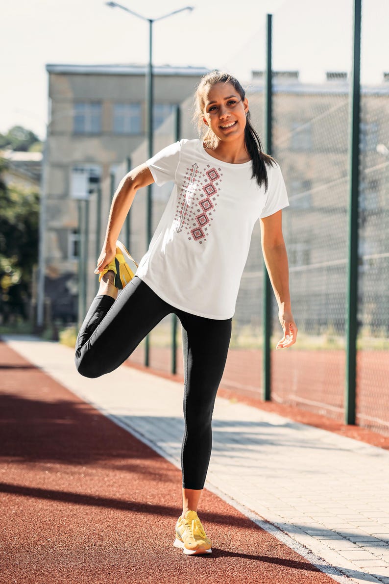 Puede incluir: Camiseta blanca con un dise&ntilde;o geom&eacute;trico en rojo y gris. La camiseta se combina con leggings negros y zapatillas deportivas amarillas. La imagen est&aacute; tomada al aire libre en una pista roja.