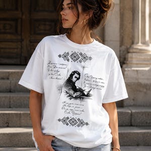 May include: White t-shirt featuring a black and white illustration of a man writing, with text in a foreign language. The shirt has decorative patterns and text in a script. The model is wearing blue jeans.