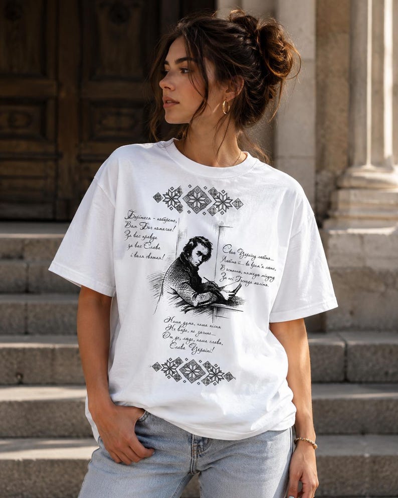 May include: White t-shirt featuring a black and white illustration of a man writing, with text in a foreign language. The shirt has decorative patterns and text in a script. The model is wearing blue jeans.