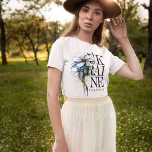 Peut inclure: Une femme portant un t-shirt blanc avec un motif de fleur bleu et blanc et le texte "UKRAINE FREEDOM". Elle porte &eacute;galement un chapeau marron et une jupe blanche.