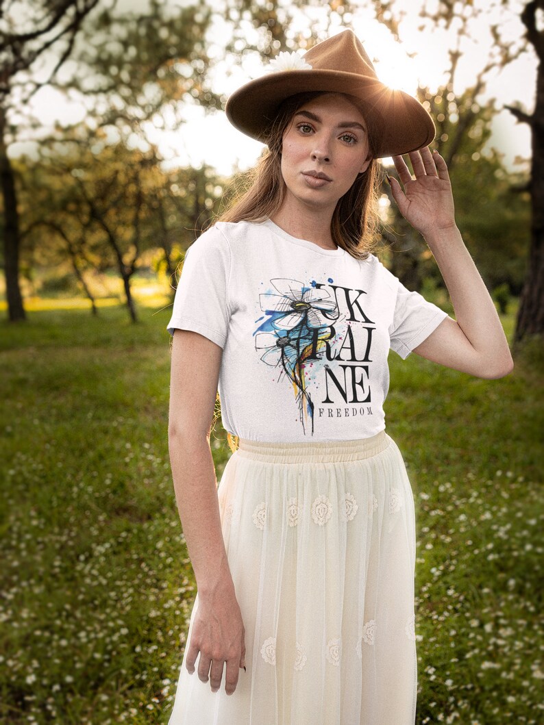 Peut inclure: Une femme portant un t-shirt blanc avec un motif de fleur bleu et blanc et le texte "UKRAINE FREEDOM". Elle porte &eacute;galement un chapeau marron et une jupe blanche.