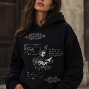 May include: Black hoodie with a hood and long sleeves. The front features a sketch of a man and script in a foreign language, along with decorative geometric patterns. The hoodie is worn by a person.