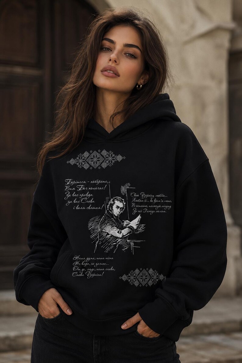 May include: Black hoodie with a hood and long sleeves. The front features a sketch of a man and script in a foreign language, along with decorative geometric patterns. The hoodie is worn by a person.