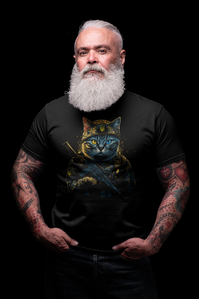 以下が含まれることがあります： 黒いTシャツ。軍服姿の猫がライフルを持っている詳細なイラスト。猫は片目が黄色、もう片方が青色で、ウクライナの三叉槍のヘルメットをかぶっています。デザインは黒い背景にあります。