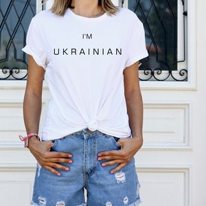 K&ouml;nnte beinhalten: Ein wei&szlig;es T-Shirt mit dem Aufdruck "I'm Ukrainian" auf der Vorderseite. Das Shirt ist in der Taille geknotet und die Person, die es tr&auml;gt, tr&auml;gt auch eine blaue Jeans-Shorts.