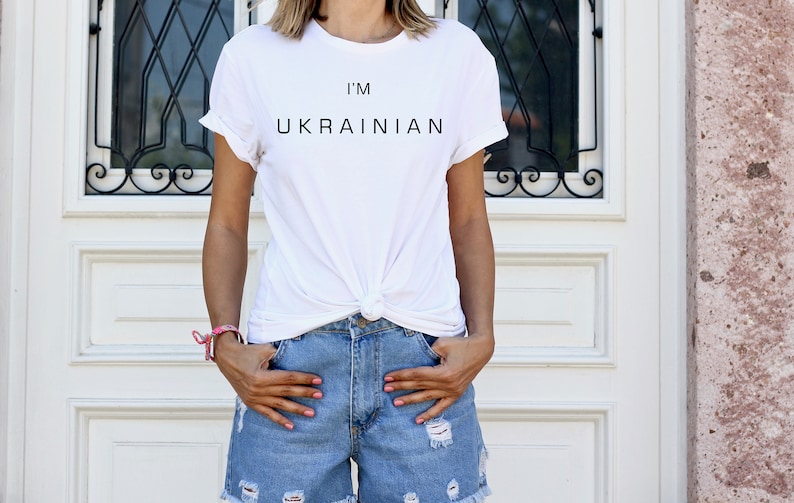 K&ouml;nnte beinhalten: Ein wei&szlig;es T-Shirt mit dem Aufdruck "I'm Ukrainian" auf der Vorderseite. Das Shirt ist in der Taille geknotet und die Person, die es tr&auml;gt, tr&auml;gt auch eine blaue Jeans-Shorts.