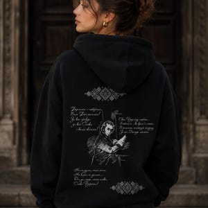 May include: Black hoodie with a detailed graphic on the back. The design includes a portrait, text, and decorative patterns in a light gray color. The hoodie is worn by a person with dark hair.