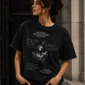 May include: Black t-shirt featuring a sketch of a person writing, with text in a foreign language. The shirt has a vintage, worn look. Decorative geometric patterns are printed above and below the illustration.