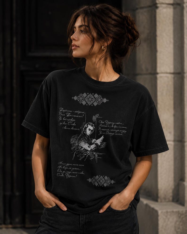 May include: Black t-shirt featuring a sketch of a person writing, with text in a foreign language. The shirt has a vintage, worn look. Decorative geometric patterns are printed above and below the illustration.