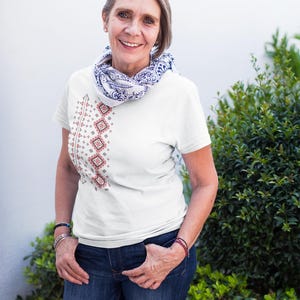 Puede incluir: Camiseta blanca con un dise&ntilde;o bordado vertical en rojo y marr&oacute;n. El dise&ntilde;o est&aacute; en el lado izquierdo de la camiseta. La persona lleva vaqueros azules y una bufanda con estampado azul y blanco. El fondo incluye plantas verdes y una pared blanca.