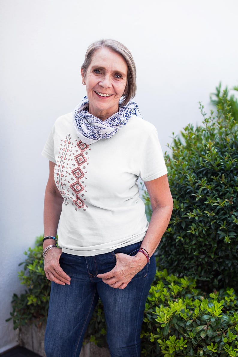 Puede incluir: Camiseta blanca con un dise&ntilde;o bordado vertical en rojo y marr&oacute;n. El dise&ntilde;o est&aacute; en el lado izquierdo de la camiseta. La persona lleva vaqueros azules y una bufanda con estampado azul y blanco. El fondo incluye plantas verdes y una pared blanca.