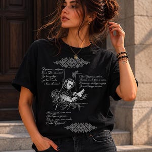 May include: Black t-shirt featuring a detailed white illustration of a man writing, with text in a foreign language. The shirt also has decorative patterns and additional text. The shirt is worn by a person.
