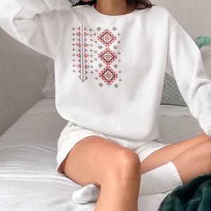May include: White sweatshirt with a red and black geometric pattern. The pattern is a traditional design with repeating squares and diamonds.