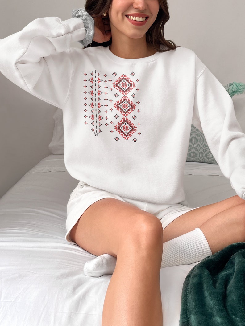 May include: White sweatshirt with a red and black geometric pattern. The pattern is a traditional design with repeating squares and diamonds.
