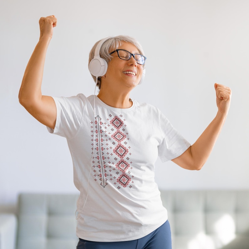 Puede incluir: Una camiseta blanca con un dise&ntilde;o geom&eacute;trico vertical en rojo y gris. La persona lleva auriculares blancos y gafas. La persona sonr&iacute;e con los brazos en alto.