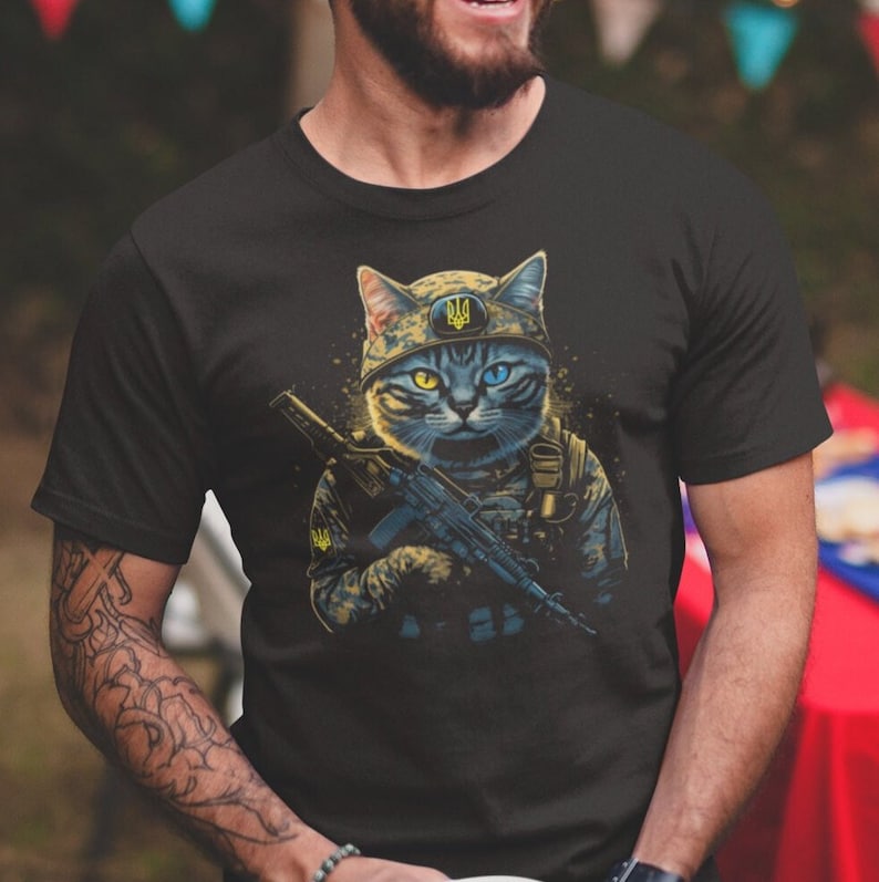 以下が含まれることがあります： 黒いTシャツ。軍服を着てライフルを持っている猫の詳細なイラスト。猫は黄色と青の目を持ち、紋章の付いたヘルメットをかぶっています。デザインはぼやけた背景にあります。