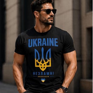Op de afbeelding: Zwart t-shirt met het woord "UKRAINE" in blauw, een blauw en geel drietandsymbool en het Oekra&iuml;ense woord "НЕЗЛАМНІ" (Onbreekbaar). Het shirt heeft ook een klein blauw en geel hartsymbool.