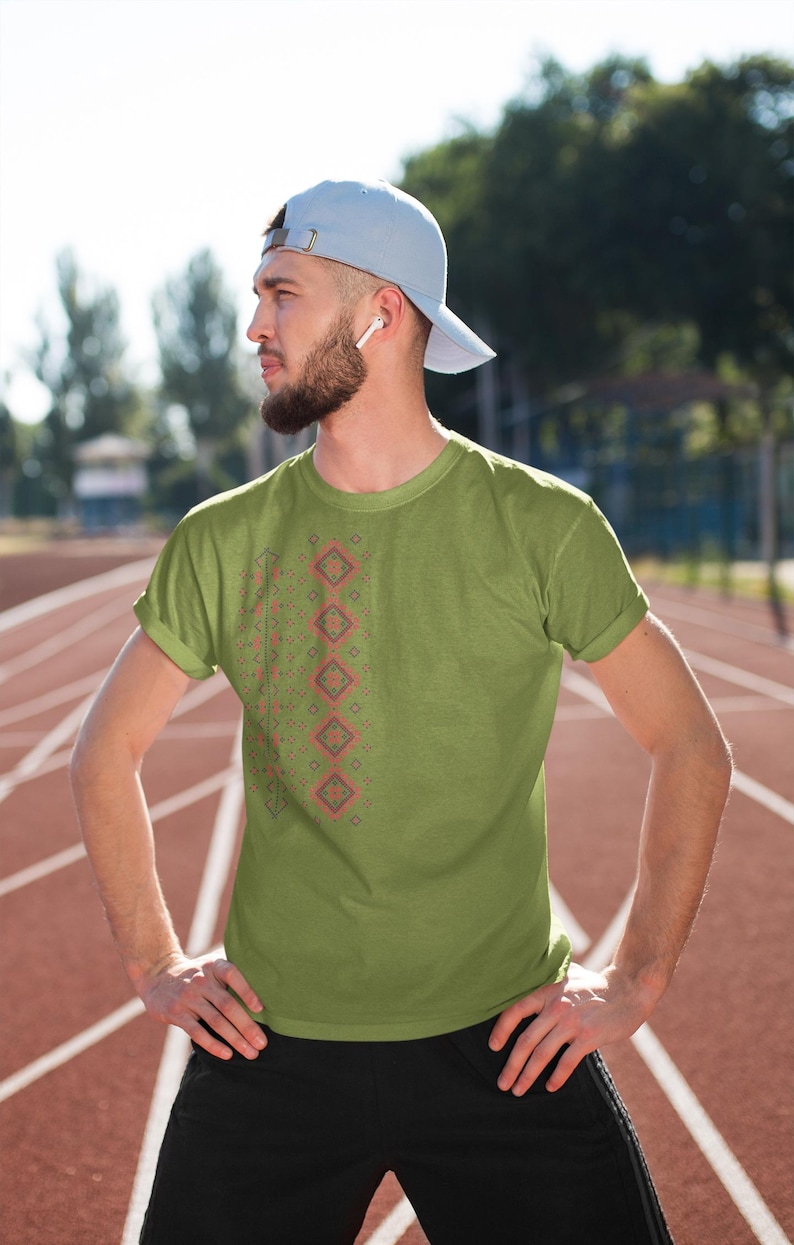 Puede incluir: Camiseta verde oliva con un patr&oacute;n geom&eacute;trico en rojo y gris. La camiseta se usa con pantalones deportivos negros y una gorra de b&eacute;isbol azul claro. La persona est&aacute; de pie en una pista roja.