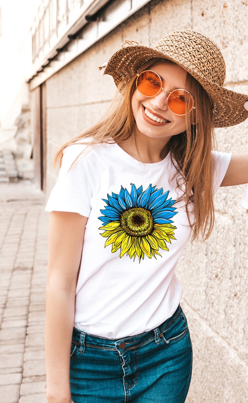 May include: A white t-shirt with a sunflower graphic. The sunflower is yellow and has blue petals. The sunflower is split in half, with the top half being blue and the bottom half being yellow.