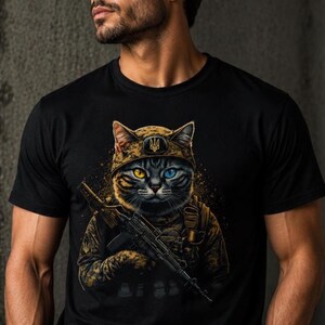 以下が含まれることがあります： 黒いTシャツ。ウクライナの三叉槍のエンブレムが付いた迷彩ヘルメットをかぶった軍服姿の猫の精巧なイラスト。猫は片目が黄色、もう片方が青色で、ライフルを持っています。デザインは金色のアクセントで詳細に描かれています。