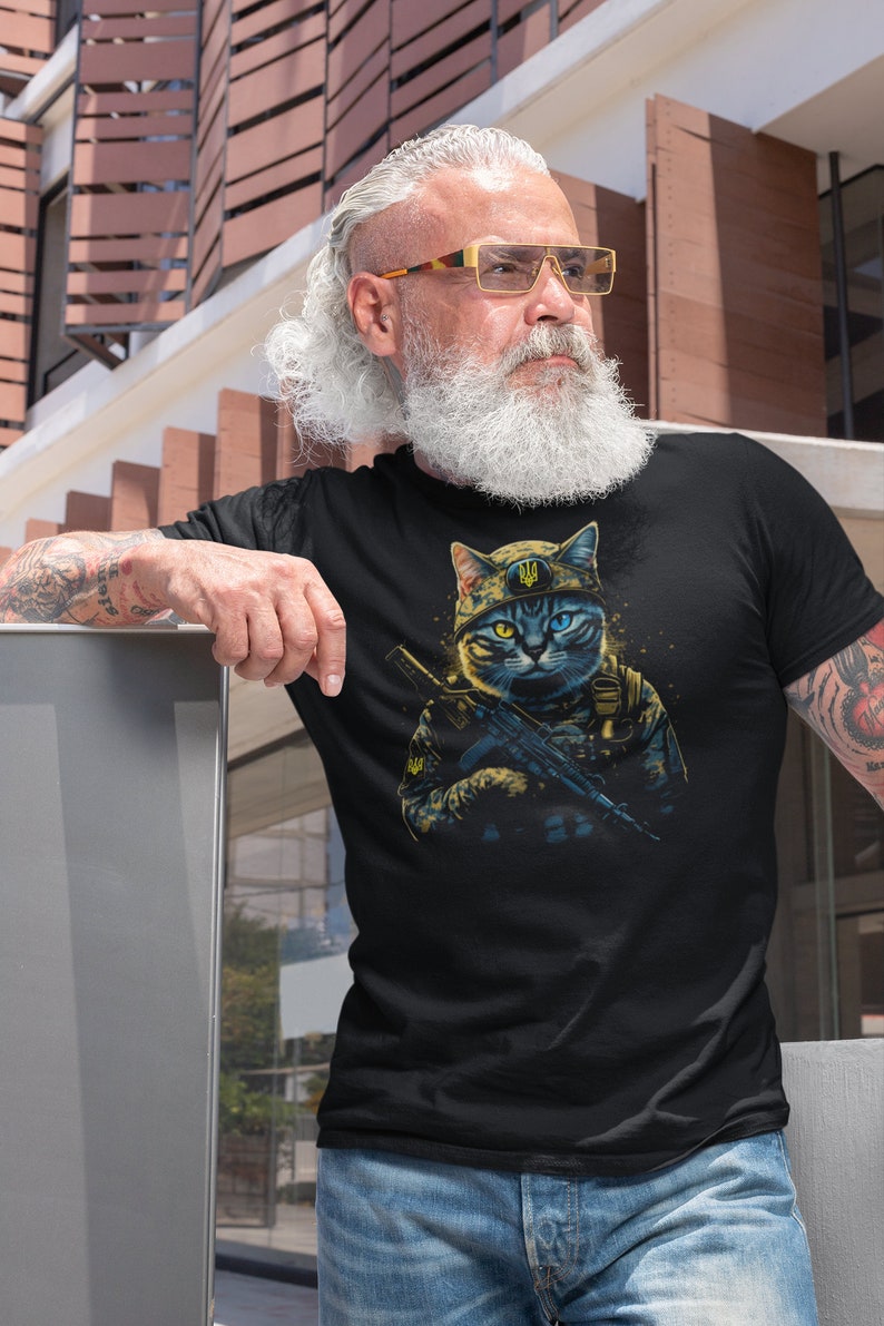 以下が含まれることがあります： 黒いTシャツ。軍服を着てライフルを持っている猫の詳細なイラスト。猫は黄色と青の目を持ち、紋章が付いた軍用帽子をかぶっています。デザインは黒い背景にあります。