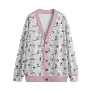 Op de afbeelding: Een vest met lange mouwen en een V-hals met een herhalend patroon van kleine wasberen, pastelkleurige regenbogen en kleine bloemetjes op een witte achtergrond. Het kledingstuk heeft een effen roze bies langs de opening, boorden en zoom.