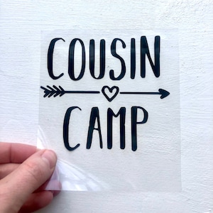 May include: Black vinyl sticker with the text "COUSIN CAMP" in a handwritten font. An arrow with a heart in the middle is between the words.