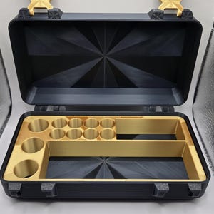 May include: A black rectangular storage case with a hinged lid and gold-colored latches. The interior features a gold-colored insert with multiple circular compartments and two rectangular sections, set against a black geometric base.