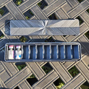 May include: A black rectangular storage case with a geometric starburst pattern on the lid. The open case features a series of vertical slots, with two small glass vials with pink and blue caps placed in the first two compartments on the left.