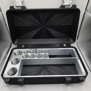 May include: A black rectangular storage case with a hinged lid featuring a sunburst pattern. The interior contains a silver-colored insert with eight circular compartments and two rectangular sections for organizing small items or components.