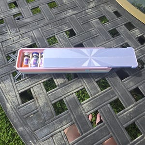 Könnte beinhalten: Ein rechteckiges, pastellfarbenes Schiebe-Etui mit einem hellvioletten Deckel, der ein weißes Sternenmuster aufweist. Das Etui ist teilweise geöffnet und gibt den Blick auf kleine Glasfläschchen mit bunten Etiketten im Inneren frei.