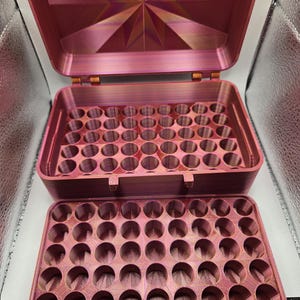 May include: A rectangular, metallic pink storage case with a hinged lid featuring a starburst pattern. The interior of the case and the removable top tray contain a grid of circular holes for organizing small items, with a visible layered texture.