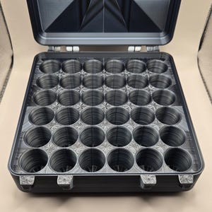 May include: A dark gray, square storage case with an open lid featuring a geometric star pattern. The interior contains a grid of thirty-six circular compartments arranged in a six-by-six layout, designed for holding small cylindrical items.