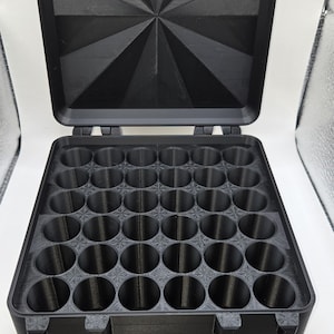 May include: A black, rectangular storage case with an open lid featuring a geometric, starburst-like pattern. The interior base contains a grid of thirty-six circular compartments arranged in a six-by-six layout, designed for organizing small items.