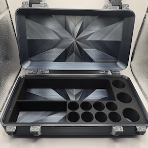 May include: A rectangular black storage case with a hinged lid, featuring a geometric starburst pattern on the interior surfaces. The base contains a black foam insert with one large rectangular compartment and twelve circular cutouts of varying sizes.