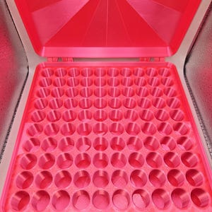 May include: A bright pink storage case with a hinged lid featuring a geometric starburst pattern. The interior contains a grid of eighty-one circular compartments arranged in a nine-by-nine square formation, designed for organizing small items.