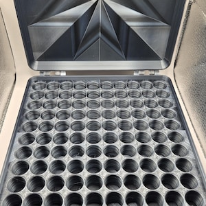 May include: A dark gray, rectangular storage case with a hinged lid. The interior features a grid of one hundred circular compartments for small items. The underside of the lid displays a geometric, star-like pattern with radiating lines.