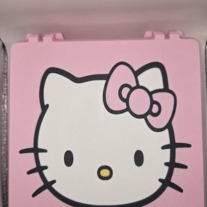 May include: A square, light pink compact case featuring a graphic of the Hello Kitty character on the lid. The character has a white face, black eyes, a yellow nose, and a light pink bow, all outlined in bold black lines.