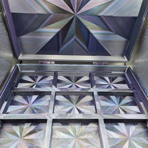 May include: A square, hinged storage box featuring a nine-compartment grid interior. Both the lid and the base of each compartment display a geometric, starburst-like pattern with a metallic, iridescent finish shifting between shades of purple, blue, and silver.