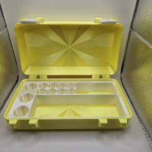 May include: A bright yellow rectangular storage case with a hinged lid featuring a sunburst pattern. The interior contains a white plastic insert with multiple circular compartments and two rectangular slots for organizing small items.