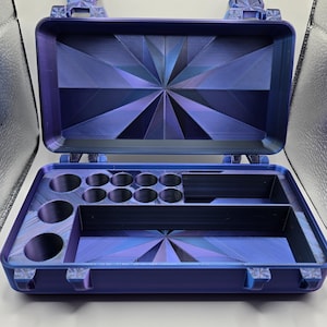 May include: A rectangular, open storage case in a deep purple and blue iridescent finish. The interior lid features a geometric starburst pattern. The base contains multiple circular compartments of varying sizes and two rectangular storage sections.