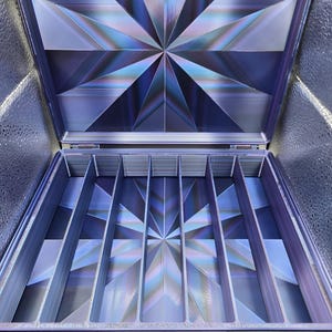 May include: A rectangular storage case with a hinged lid, featuring a vibrant purple and blue starburst pattern on the interior base and lid. The interior is divided into six long, narrow vertical compartments by five thin, upright dividers.