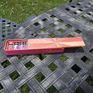 Könnte beinhalten: Ein rechteckiges, rosafarbenes Schiebegehäuse mit vier kleinen Glasfläschchen mit blauen Verschlüssen. Der Schiebedeckel zeigt ein geometrisches Strahlenmuster in Orange-, Rosa- und Gelbtönen. Das Gehäuse ist mit einem seitlichen Kerbmechanismus versehen.