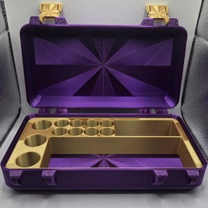 May include: A purple storage case with a hinged lid and gold-colored latches. The interior features a gold-colored insert with multiple circular compartments and two rectangular sections, set against a purple base with a geometric sunburst pattern.