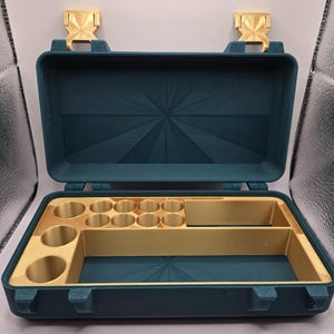 May include: A teal rectangular storage case with a hinged lid and gold-colored latches. The interior features a gold-colored insert with multiple circular compartments and two rectangular sections, set against a dark teal base with a sunburst pattern.