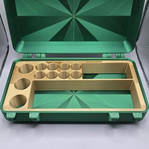 May include: A rectangular green storage case with a hinged lid featuring a sunburst pattern. The interior contains a tan insert with ten circular compartments and two rectangular slots for organized storage, secured by two light-colored latches.