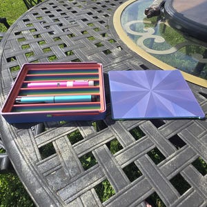 Könnte beinhalten: Ein rechteckiges violettes Aufbewahrungstablett mit mehrfarbigem Innenraum und horizontalen Schlitzen, in denen ein rosa und ein hellblauer Stift liegen, sowie ein separater quadratischer violetter Deckel mit geometrischem Sternmuster.