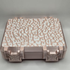 May include: A rectangular storage case with a rose gold metallic finish and two front latches. The top surface features a white and rose gold leopard print pattern with a textured appearance. The case has a structured, boxy design.